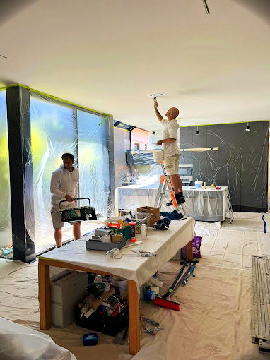 Professional painter standing on a table to reach the ceiling in a modern interior with large windows, illustrating the safe and thorough painting process.
